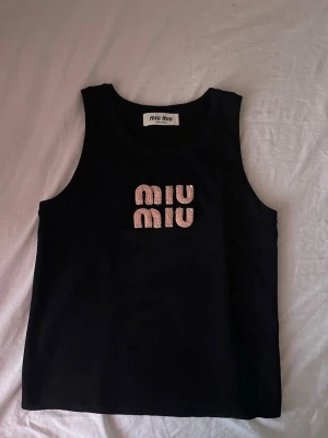 Svart linnetopp från Miu Miu - Snygg svart linnetopp från Miu Miu med rosa pärlbroderat logotyp på bröstet. Toppen är ärmlös och har en klassisk rund halsringning. Perfekt för dig som vill ha en trendig och stilren topp med exklusiv känsla.