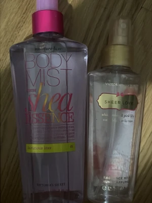 Victoria's Secret Body mists old bottles  - Två fräscha body mists från Victoria's Secret: 'Sheer Love' med vit bomullsblomma och rosa lilja, samt 'Luminous Kiss' med shea essence. Båda i genomskinliga plastflaskor, en med rosa och guld detaljer och den andra med guld och vit etikett.