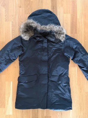 Svart parkas från Didriksons, 100% vattentätt (StormSystem) - Stilren svart parkas från Didriksons med huva och avtagbar fuskpäls. Jackan har två stora fickor framtill med lock och knappar samt dragkedja och vindslå. Perfekt för kalla dagar med sin långa modell och skyddande material. 100% vattentätt med Storsystem.  