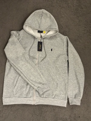 Grå hoodie från Polo Ralph Lauren - Snygg ljusgrå hoodie med dragkedja från Polo Ralph Lauren. Klassisk design med svart broderad logga på bröstet, huva med dragsko och två fickor framtill. Mjuk bomullsblandning som är skön att ha på sig. Perfekt för en avslappnad och stilren look.