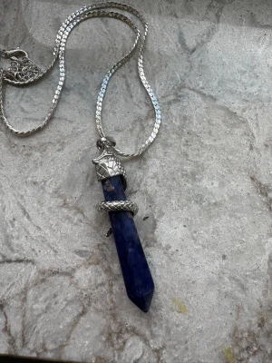 Halsband med sodalit kristall och orm - Unikt halsband med en spetsig sodalit kristall som hänge, dekorerad med en silverfärgad orm som slingrar sig runt stenen. Kedjan är silverfärgad och har ett diskret mönster. Perfekt för dig som gillar mystiska och coola accessoarer.