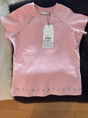 Rosa t-shirt med stjärndetaljer, helt ny och oanvänd  - Snygg rosa t-shirt från Zara med silverfärgade stjärnor och nitar längs axlar och nederkant. T-shirten har korta ärmar och en klassisk rund halsringning. 