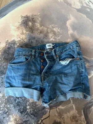 Blå jeansshorts från 157, XS - Snygga blå jeansshorts från 157 i storlek XS. Klassisk femficksmodell med uppvikta benslut och knappar framtill. Perfekta till sommaren och lätta att matcha med allt. Tillverkade i bomull för en skön känsla.