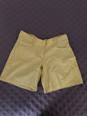 Gula shorts med knappar - Snygga gula shorts med flera dekorativa knappar och fickor både fram och bak. Klassisk passform med bälteshällor och tillverkade i ett mjukt bomullstyg. Perfekta för varma dagar och enkel att matcha med olika toppar.