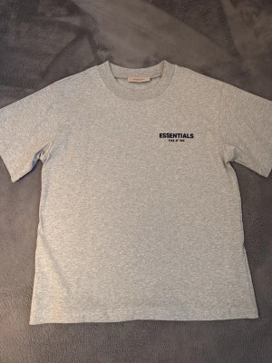 Grå Essentials T-shirt Fear of God - Snygg grå T-shirt från Fear of God Essentials med svart logga tryckt på bröstet och ryggen. Klassisk rund hals och korta ärmar. Perfekt för en clean och stilren look. Gjord i mjuk bomull för maximal komfort.