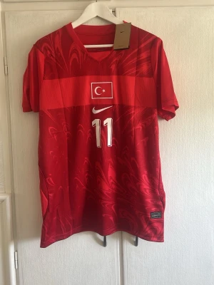 Turkiet fotbollströja Nike röd #11 yildiz - Röd officiell Turkiet fotbollströja från Nike med nummer 11 och namnet Yildiz på ryggen. Tröjan har korta ärmar, V-ringning och Turkiets flagga på bröstet. Perfekt för dig som vill stötta landslaget med stil.
