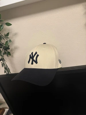 New Era Yankees beige keps - Snygg beige keps från New Era med mörkblått NY Yankees-broderi framtill och mörkblå skärm. Klassisk böjd skärm och justerbar passform bak. Perfekt för dig som vill ha en stilren och sportig look. Den är iprincip sprillansny