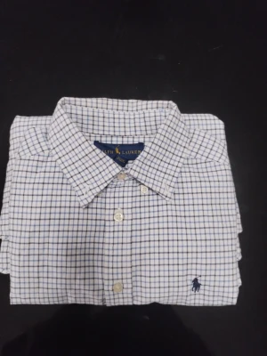 Rutig skjorta från Ralph Lauren - Klassisk vit skjorta från Ralph Lauren med blått rutmönster och broderad logga på bröstet. Skjortan har knappar framtill och krage, perfekt för en stilren look. Tillverkad i mjuk bomull som känns skön mot huden.