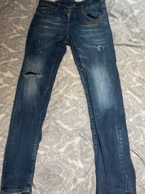 Dsquared2 jeans mörk blåa  - Säljer ett par blå dsquared2 jeans med slitningar, hål på ena knät och coola färgstänk. Jeansen har en klassisk femficksmodell och raka ben. Perfekta för en avslappnad och trendig look.