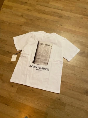 Vit t-shirt med tryck från Aimé Leon Dore - Vit t-shirt från Aimé Leon Dore med svart tryck på både fram- och baksida. På ryggen finns ett stort tidningsinspirerat motiv och texten 'A FAMILY BUSINESS.' samt 'THE WORLD'S BOROUGH' på bröstet. Klassisk passform och rund hals. Perfekt för en clean och stilren look.