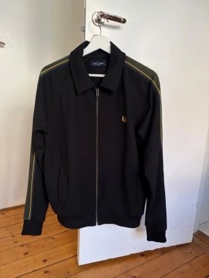 Svart track jacket från Fred Perry - Svart zip tröja från fred Perry med gröna och gula detaljer. Ser ut som ny och kan skippa mått om det behövs. 
