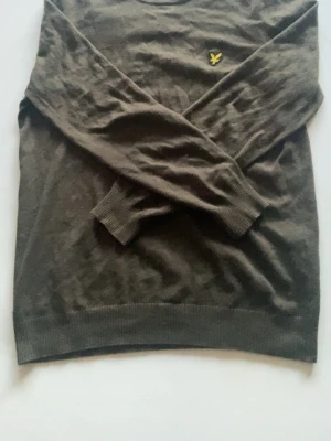 Olivgrön stickad tröja Lyle & Scott - Olivgrön stickad tröja från Lyle & Scott med rund halsringning och klassisk gul logga broderad på bröstet. Tröjan har ribbade muddar vid ärmslut och nederkant. Perfekt för lager-på-lager och enkel att matcha med jeans eller chinos.