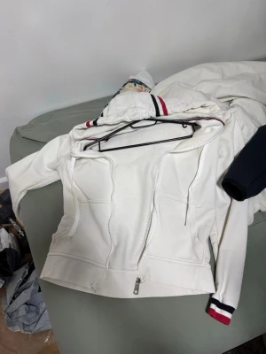 Vit Moncler hoodie med dragkedja XS - Snygg vit hoodie från Moncler i storlek XS med en trasig dragkedja framtill. Detaljer i rött och marinblått på huva och ärmslut ger en sportig vibe. Tillverkad i mjukt material och har två fickor framtill. Perfekt för en chill och stilren look.