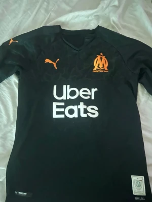 Svart Marseille matchtröja Puma - Svart fotbollströja från Puma med Marseille-logga i orange och Uber Eats-tryck på bröstet. Tröjan har korta ärmar, V-ringning och är tillverkad i funktionsmaterial med DryCell-teknologi. Perfekt för träning eller match.