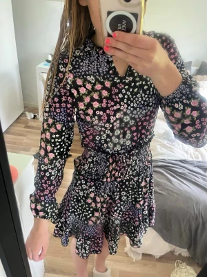 Svart blommig kortklänning - Supersöt svart kortklänning med färgglatt blommönster i rosa, lila, vitt och grönt. Klänningen har långärmade ärmar, markerad midja med smock och volangig kjol. Perfekt för dig som gillar en feminin och lekfull stil.