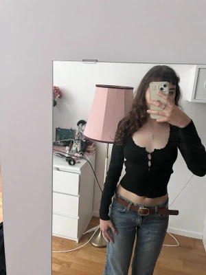 Svart croppad topp med knappar - Snygg svart croppad topp med lång ärm och djup v-ringning. Toppen har dekorativa knappar framtill och vågig kant nertill. Figurnära passform som framhäver midjan. Perfekt att styla med jeans eller kjol.