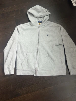 Grå hoodie från Polo Ralph Lauren - Snygg grå hoodie från Polo Ralph Lauren med dragkedja, huva och två fickor framtill. Klassisk broderad logga på bröstet och mjukt material som känns skönt mot huden. Perfekt för en avslappnad och stilren look.