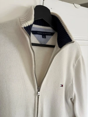 Vit ziptröja från Tommy Hilfiger - Vit stickad ziptröja från Tommy Hilfiger med ribbad hög krage och mörkblå insida. Tröjan har dragkedja framtill och diskret logga broderad på bröstet. Perfekt för en stilren och avslappnad look.