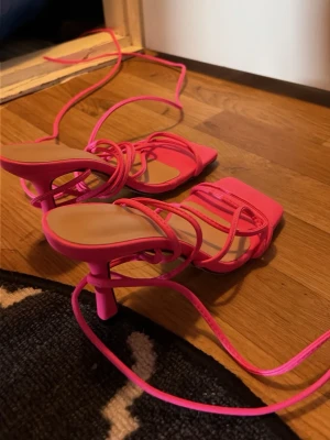 Rosa sandaletter med snörning - Säljer ett par trendiga sandaletter i starkt rosa färg med tunna remmar och snörning som går att vira runt benet. Skorna har fyrkantig tå och medelhög klack. Perfekta för dig som vill sticka ut och addera färg till din outfit.