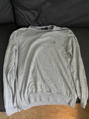 Grå tröja från Polo Ralph Lauren - Grå långärmad tröja från Polo Ralph Lauren i storlek M med klassisk broderad logga på bröstet. Tröjan har rund halsringning och ribbade muddar vid ärmslut och nederkant. Perfekt för en stilren och enkel look.