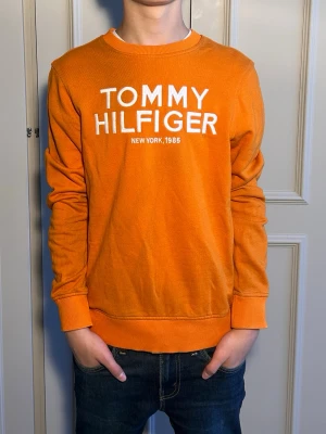 Orange sweatshirt Tommy Hilfiger - Snygg orange sweatshirt från Tommy Hilfiger med vit logotyptext framtill och rund halsringning. Tröjan har ribbade muddar vid ärmslut och nederkant samt en avslappnad passform. Perfekt för en casual och trendig look. Skriv ifall ni har några frågot