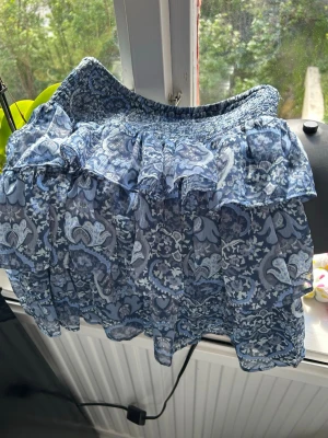 Blå blommig volangkjol från Lindex - Supersöt blå kortkjol från Lindex med blommigt mönster och volanger. Kjolen har smock i midjan för extra komfort och är gjord i återvunnen polyester. Perfekt för dig som gillar en luftig och trendig stil.