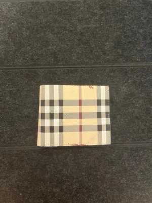 Wallet Burberry Nova Check Bi-Fold - Burberry bi-fold wallet featuring the iconic Nova Check pattern in beige, black, white, and red on the exterior, complemented by a black textured interior. The inside includes an ID window showcasing the Burberry Equestrian Knight Device logo and "ESTABLISHED 1856". Visible reddish marks are present on the exterior edges.    #