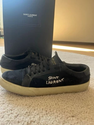 Svarta Saint Laurent sneakers - Svarta sneakers från Saint Laurent med vit handskriven logga på sidan. Skorna har lågt skaft, svarta snören och vit sula. Materialet är canvas med detaljer i mocka och läder. Klassisk och stilren design med en edgy touch.