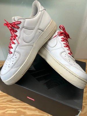 Nike Air Force 1 x Supreme - Nike Air Force 1 i samarbete med Supreme. Klassiska vita sneakers i skinn med röda Supreme-skosnören och diskret Supreme-logga på sidan. Tjock sula och ikonisk Nike Swoosh på sidorna. Perfekta för dig som gillar streetwear och vill sticka ut.