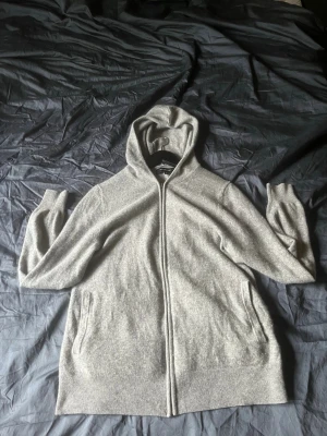 Kashmir hoodie - Mysig ljusgrå hoodie kashmir i mjukt stickat material. Hoodien har dragkedja framtill, två fickor och ribbade muddar vid ärmslut och nederkant. Perfekt för en avslappnad och stilren look.