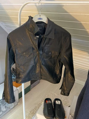 Svart cropped skinnjacka  - Snygg svart cropped skinnjacka från Fb sisters med klassisk krage, två stora bröstfickor med lock och tryckknappar framtill. Jackan har raka ärmar och en stilren, minimalistisk look som passar till många outfits.