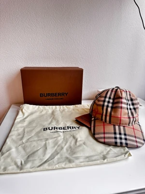 Burberry keps med klassiskt rutmönster - Säljer en snygg Burberry keps i storlek M med det ikoniska beigea rutmönstret och svarta, röda samt vita detaljer. Kepsen är tillverkad i bomull och polyester och har justerbar passform. Perfekt accessoar för att lyfta din streetwear-look.