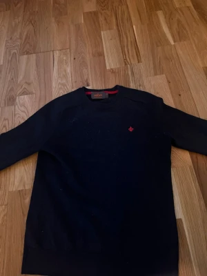 Mörkblå stickad tröja från Morris - Mörkblå stickad tröja från Morris i 100% merinoull. Tröjan har rund halsringning och en röd broderad logga på bröstet. Perfekt för dig som gillar klassisk och stilren design med en twist.