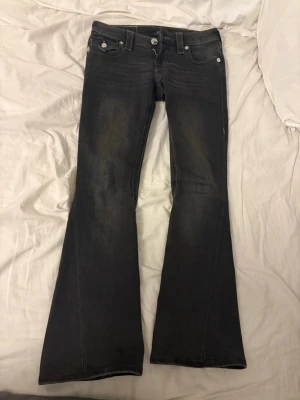 Svarta bootcut jeans från True Religion - Säljer ett par svarta bootcut jeans från True Religion med snygga kontrastsömmar och ikonisk fickdetalj bak.jeansen är 95cm långa. Jeansen har klassisk femficksdesign, låg midja och silverfärgade knappar. Perfekta för dig som gillar en retro vibe och vill sticka ut.