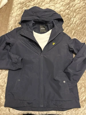 Marinblå vindjacka från Lyle & Scott - Snygg marinblå vindjacka från Lyle & Scott med huva och dragkedja framtill. Jackan har två fickor med tryckknappar, elastiska muddar vid ärmsluten och den klassiska gula loggan på bröstet. Perfekt för blåsiga dagar och enkel att matcha. Helt ny utan prislapp😃