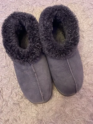 Grå fluffiga tofflor i fårskinn - Mysiga grå tofflor med fluffigt foder och ovansida i mjukt fårskinn. Tofflorna har en högre kant runt ankeln och synliga sömmar för en avslappnad look. Perfekta för att hålla fötterna varma hemma. Storleken beror på ens fötter men passar de flesta, 43-45 ish.