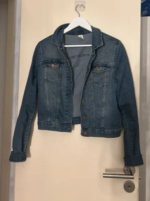 Klassisk blå jeansjacka - Snygg blå jeansjacka med raka linjer och bruna knappar framtill. Jackan har två bröstfickor med lock och knapp, samt klassisk krage. Perfekt för dig som vill ha en tidlös och trendig look.