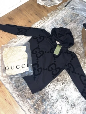 Svart hoodie med GG-mönster från Gucci - Snygg svart hoodie från Gucci med det klassiska GG-mönstret i sammet över hela tröjan. Hoodien har huva och ribbade muddar. Tillverkad i mjukt bomullsmaterial och levereras med originalpåse och tagg. Perfekt för dig som vill ha en exklusiv och trendig look.