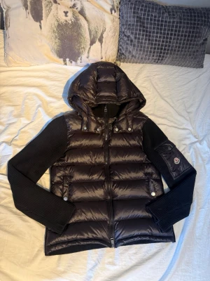 Moncler Cardigan - Snygg svart cardigan Moncler med glansig vadderad kropp och stickade ärmar. Jackan har huva, dragkedja framtill och Moncler-logga på ärmen. Perfekt för kyliga dagar och ger en sportig vibe. Skick 10/10, knappt använd. Välldigt fräsch och passar till nästan alla outfits. Passar både M-L. Storleken är 3. Påse och tags medföljer. Scan samt nfc fungerar fel fritt! Vid frågor eller intresse, tveka inte på att höra av dig! :) 