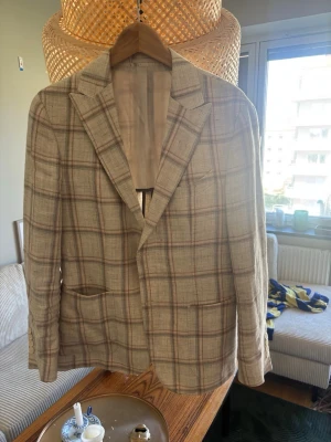 Beige rutig kavaj med klassiskt snitt - Snygg beige kavaj med rutigt mönster i brunt och grått. Kavajen har två framfickor, klassisk krage och är helfodrad. Perfekt för dig som gillar en stilren och tidlös look. Materialet känns lätt och passar bra till olika outfits.