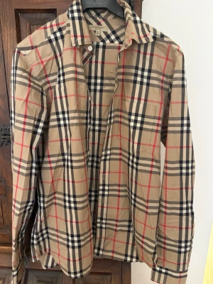 Beige rutig skjorta från Burberry - Klassisk beige skjorta från Burberry med det ikoniska rutmönstret i svart, vitt och rött. Skjortan har lång ärm, knappar framtill och en snygg krage. Perfekt för dig som vill ha en tidlös och trendig look. 
