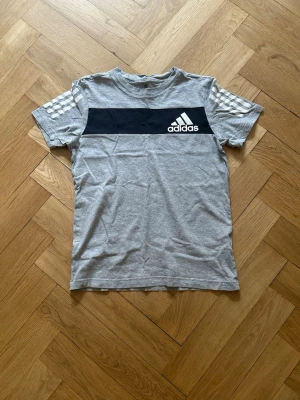  Adidas t-shirt - Snygg grå t-shirt från Adidas med svart panel och vit logga på bröstet. Klassiska vita ränder på axlarna ger en sportig vibe. Perfekt till jeans eller shorts. Tillverkad i mjuk bomull för skön känsla hela dagen.