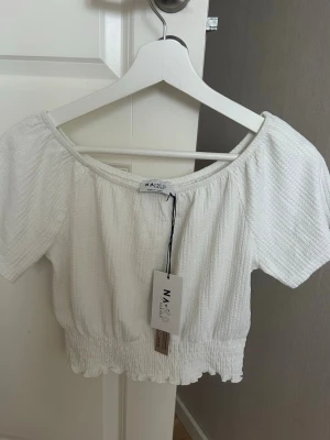 Vit offshoulder topp från NA-KD - Superfin vit offshoulder topp från NA-KD med smockad nederkant och korta ärmar. Tunt, strukturerat tyg som ger en luftig känsla. Perfekt till jeans eller kjol för en fräsch look.