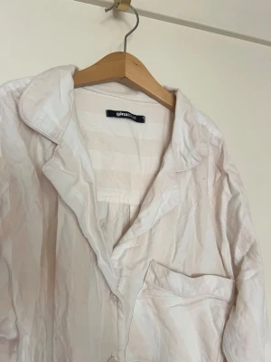 Randig pyjamasskjorta från Gina Tricot - Ljusbeige och vit randig pyjamasskjorta från Gina Tricot med klassisk krage, knäppning framtill och bröstficka. Skjortan har långa ärmar och är tillverkad i ett mjukt, lätt material som passar perfekt för mysiga hemmakvällar.