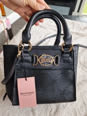 Svart handväska Juicy Couture - Snygg svart handväska från Juicy Couture med präglad logga, guldfärgade metalldetaljer och dubbla handtag. Väskan har avtagbar axelrem och innerfoder med Juicy Couture-tryck. Perfekt för dig som gillar trendiga accessoarer.