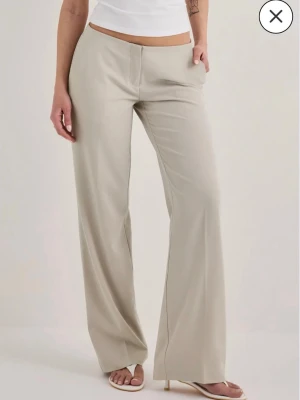Beige vida kostymbyxor - Snygga beige kostymbyxor med hög midja och vida ben. Byxorna har pressveck fram och bak samt diskreta fickor bak. Perfekta för en clean och stilren look. Tillverkade i ett mjukt, vävt material som ger en skön känsla.