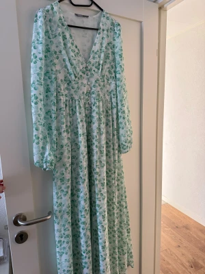 Lång blommig klänning med v-ringning - Säljer en långklänning med blommigt mönster i grönt och vitt. Klänningen har v-ringning, långa ärmar och ett luftigt fall. Perfekt för sommarens alla tillfällen. Märke: NA-KD.