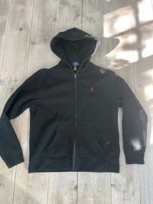 Svart hoodie från Polo Ralph Lauren - Svart hoodie med dragkedja från Polo Ralph Lauren. Klassisk design med huva, kängurufickor och röd broderad logga på bröstet. Mjuk bomullsblandning som är skön att ha på sig. Perfekt för en avslappnad och stilren look. Märkt med juniorstorlek L, 14-16 år. Skulle säga att den passar dig som är 170-180 cm lång