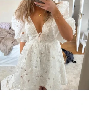 Vit blommig kortklänning med puffärm - Superfin vit kortklänning med blommigt spetsmönster, djup v-ringning och puffärmar. Klänningen har snörning framtill och är ärmlös med volangdetaljer. Perfekt för sommarens alla tillfällen!