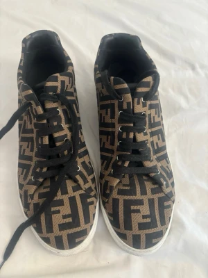 Fendi sneakers - Säljer ett par snygga sneakers från Fendi med det klassiska FF-mönstret i brunt och svart. Skorna har svarta skosnören och vit sula. .